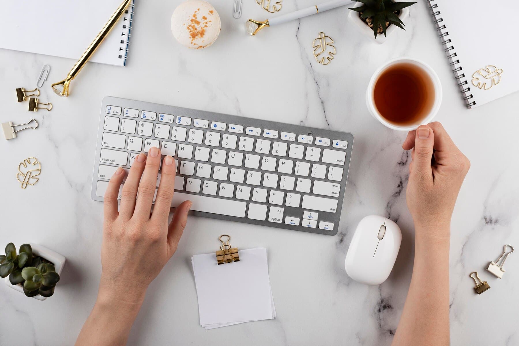 A person's hands typing on a gray keyboard, holding a cup of tea, surrounded by notebooks, stationery, and a potted plant.