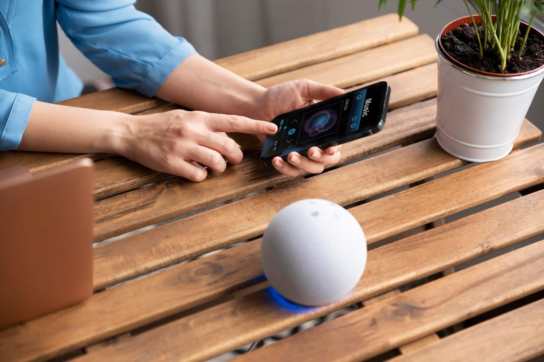 A person interacts with a smartphone app while controlling a smart speaker on a wooden table, next to a potted plant.