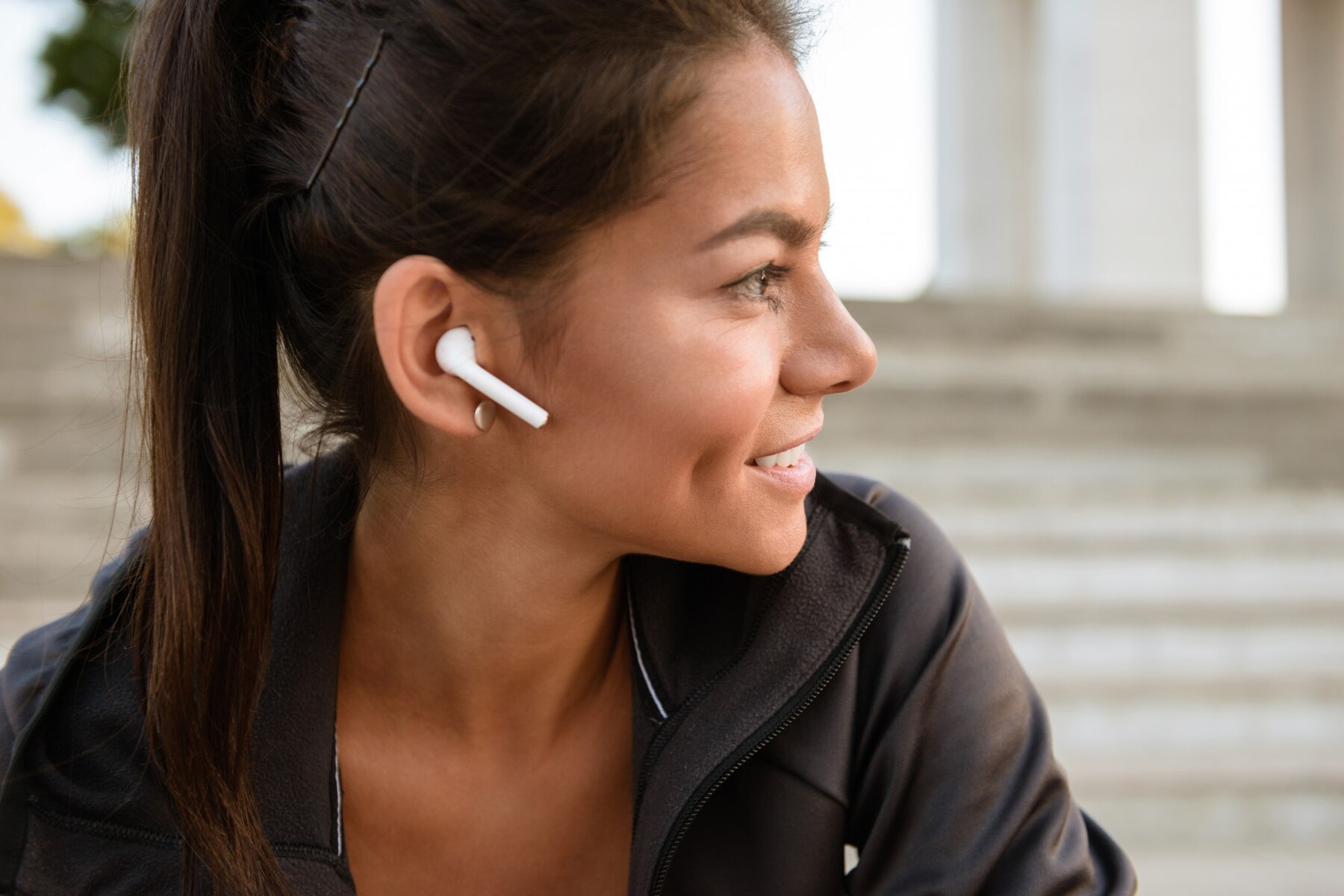 A woman with long dark hair wears white earphones while sitting outdoors, dressed in a black zip-up jacket.