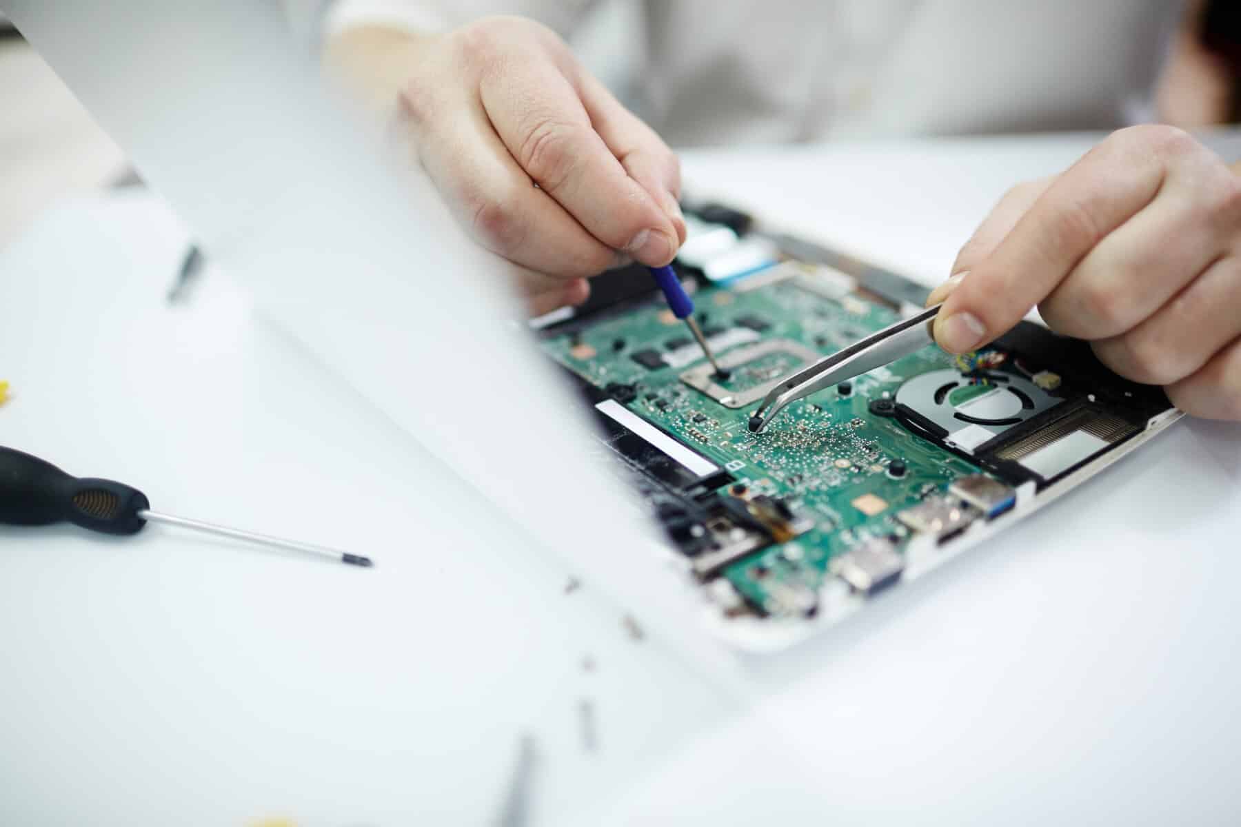 A technician is using tweezers and a screwdriver to repair a laptop's internal circuit board, focusing on a precise task at a clutter-free workstation.