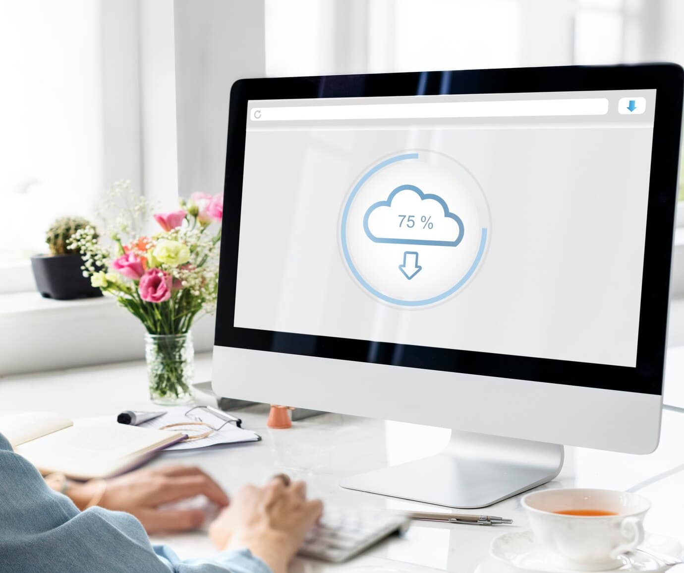 A person typing at a computer displaying a cloud download progress at 75%, with flowers and a tea cup in the background.