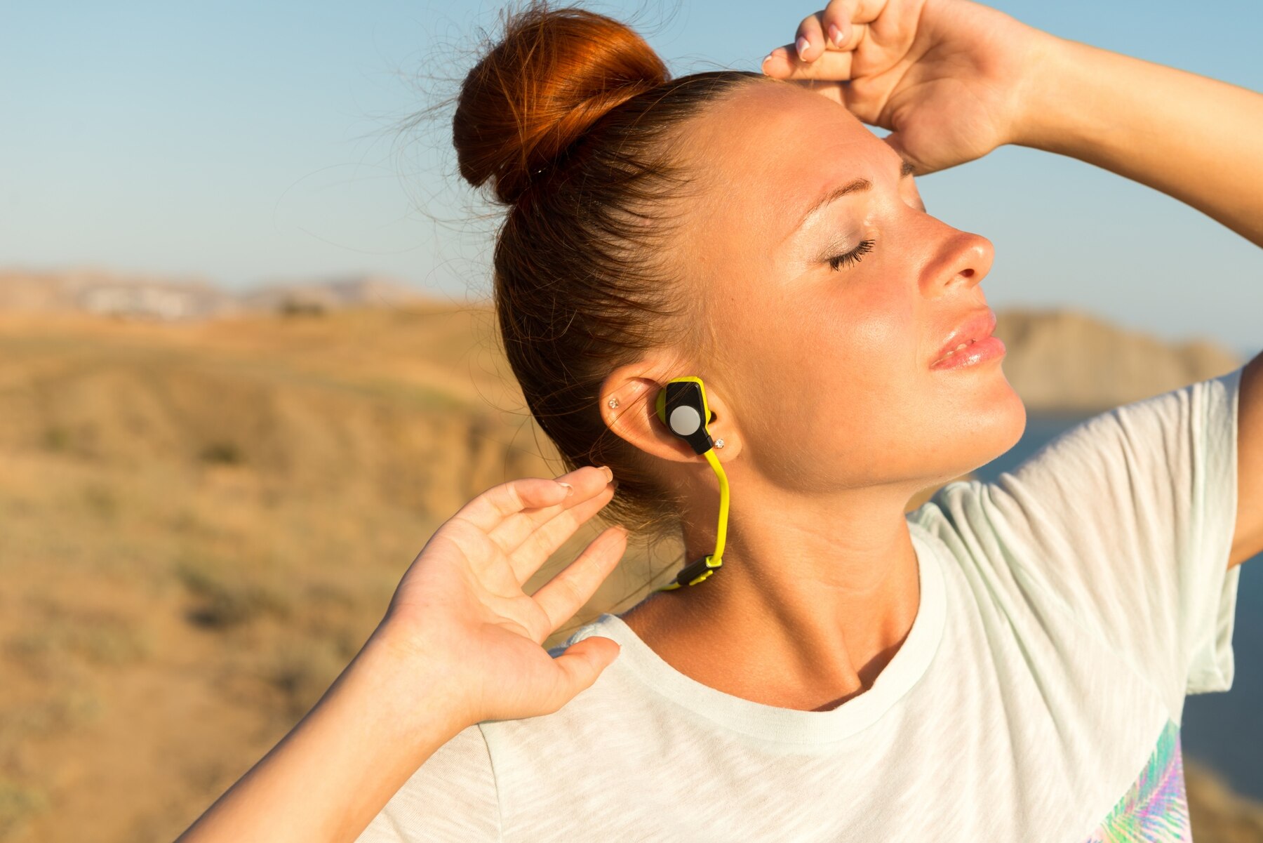 A person wearing wireless earbuds enjoys music outdoors, with a scenic backdrop of hills and clear skies.