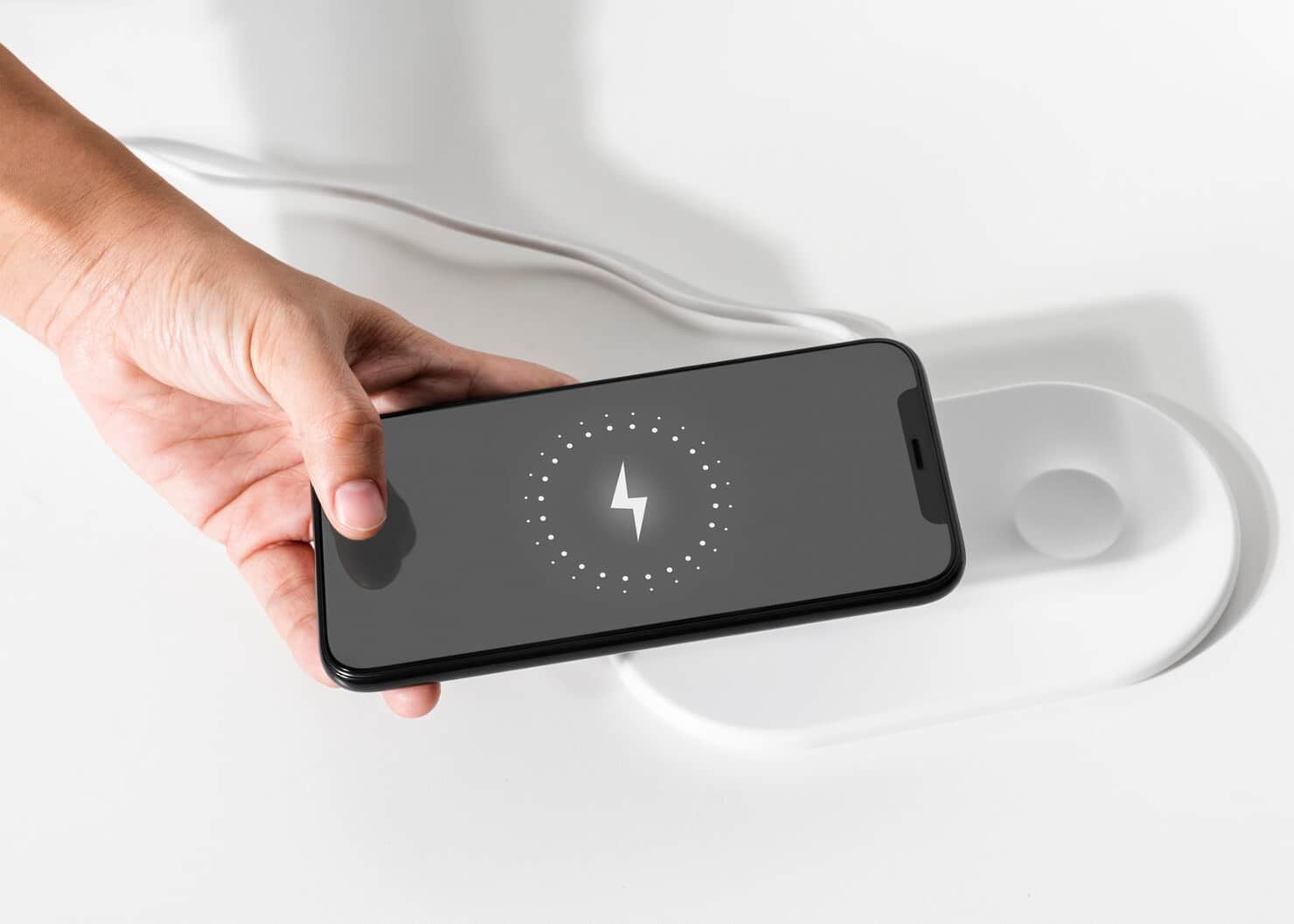 A person's hand placing a black smartphone with a charging indicator on the screen onto a white wireless charging pad.