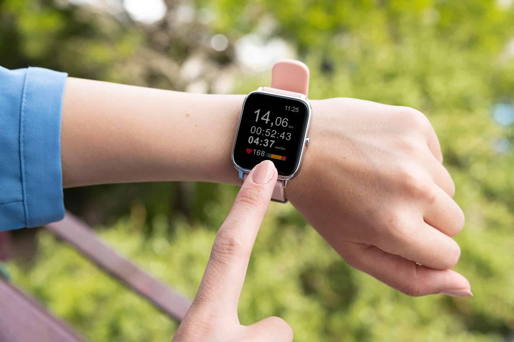 A close-up of a hand tapping a smartwatch displaying distance, time, and heart rate against a blurred green background.