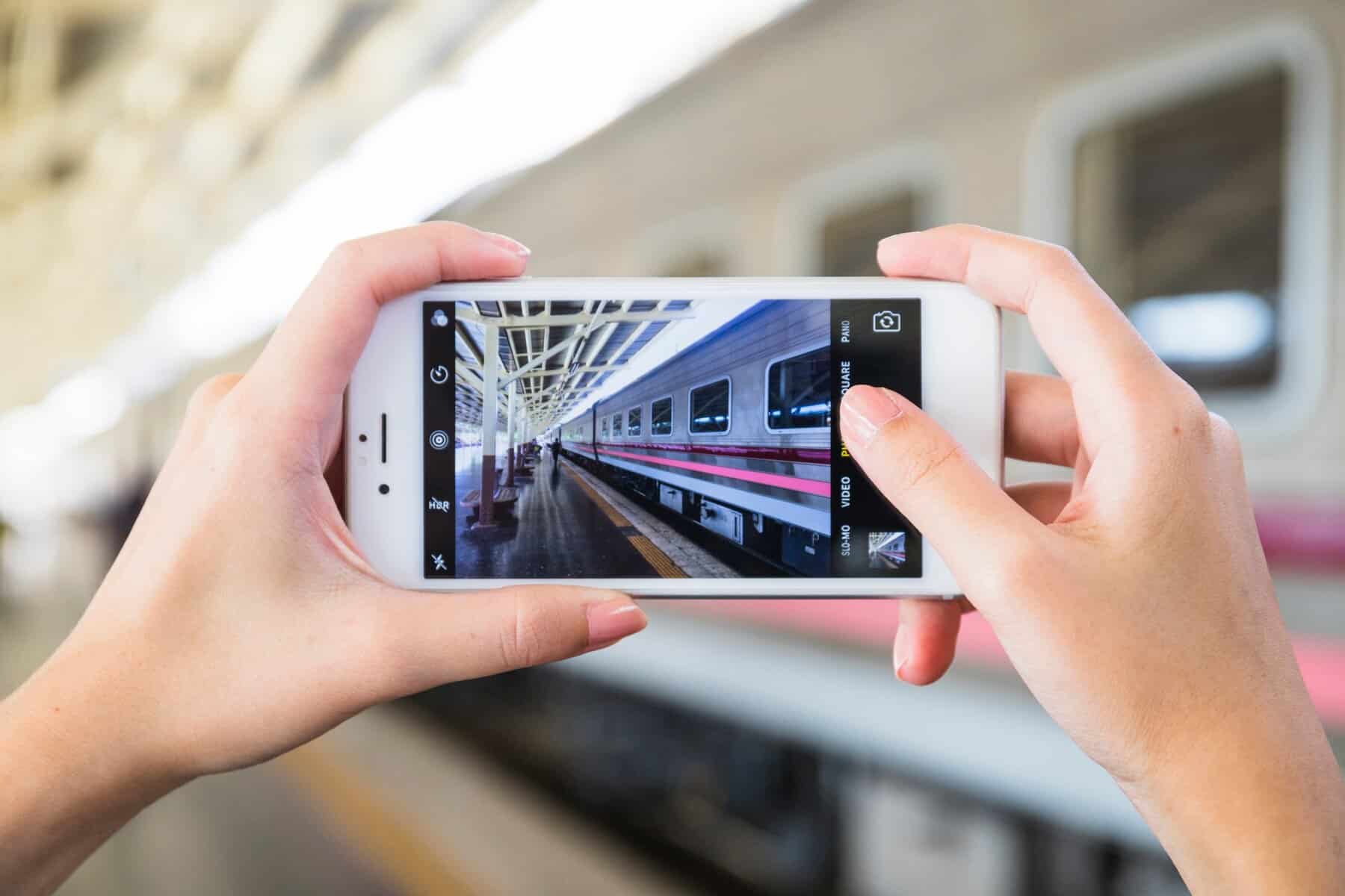 A person holds a smartphone, capturing a photo of a train at a station, with a modern architectural backdrop.