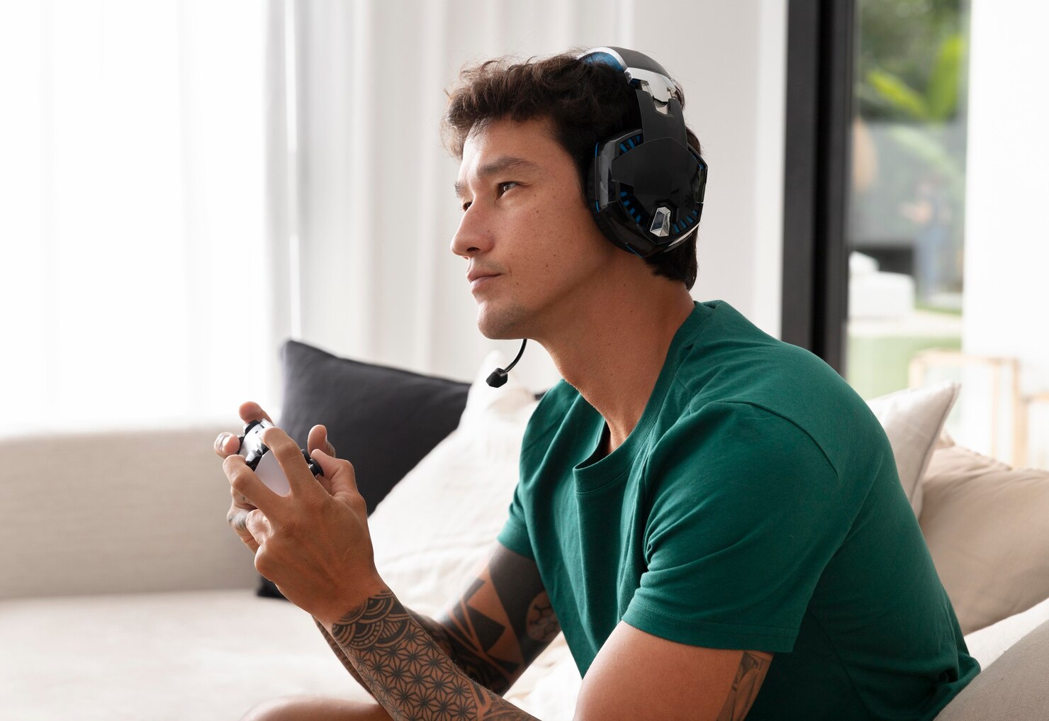 A person wearing a gaming headset holds a controller, seated on a couch in a bright room with soft natural light.