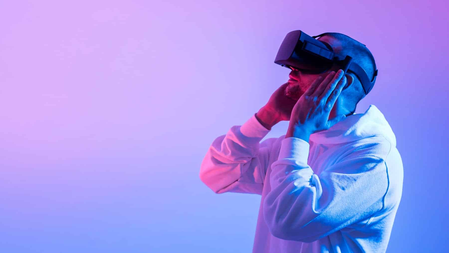 A man wearing a virtual reality headset, illuminated by pink and blue lights, touches his temple and cheek, absorbed in a digital experience.