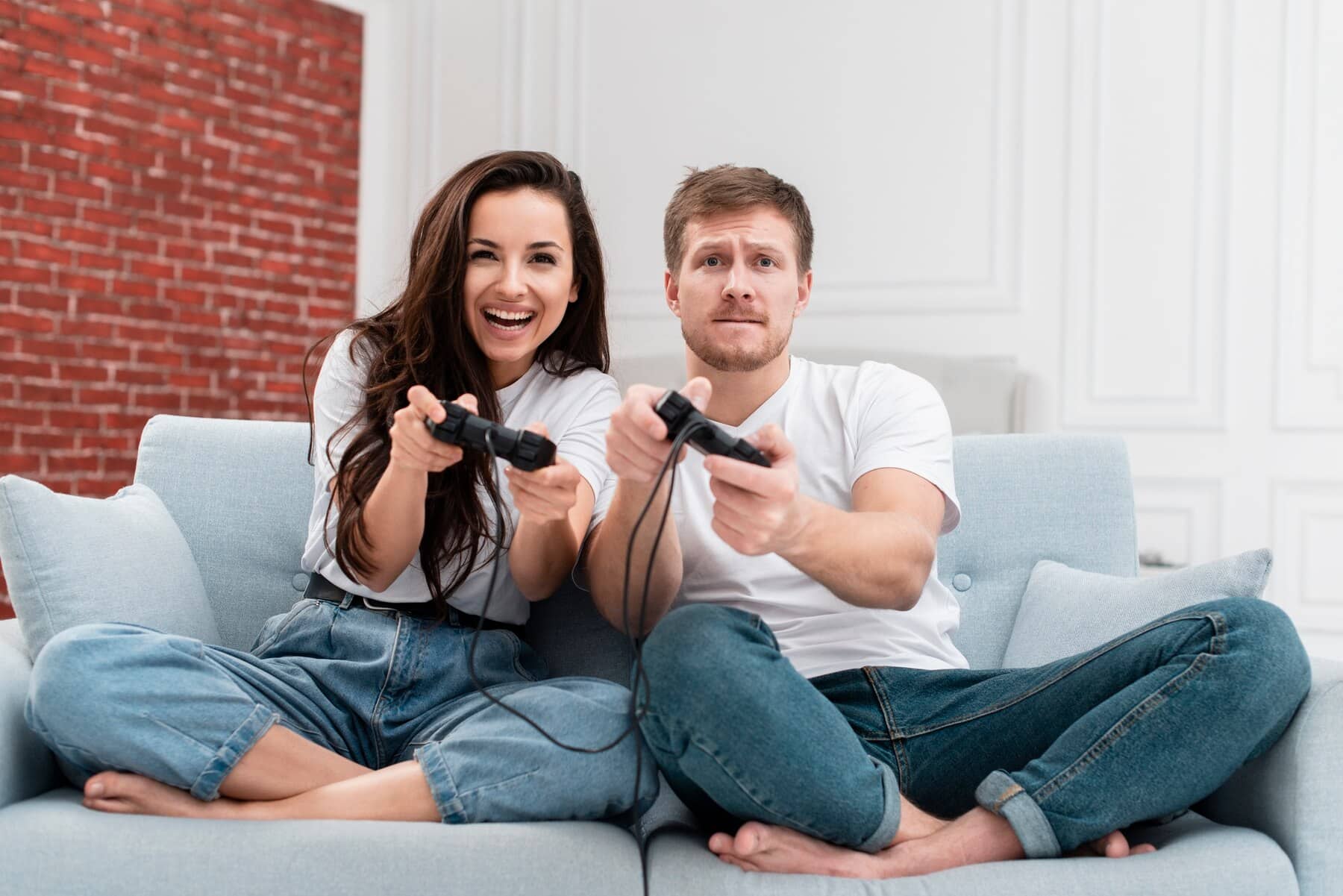 A young couple sitting on a sofa, playing video games with controllers, surrounded by a modern living room.
