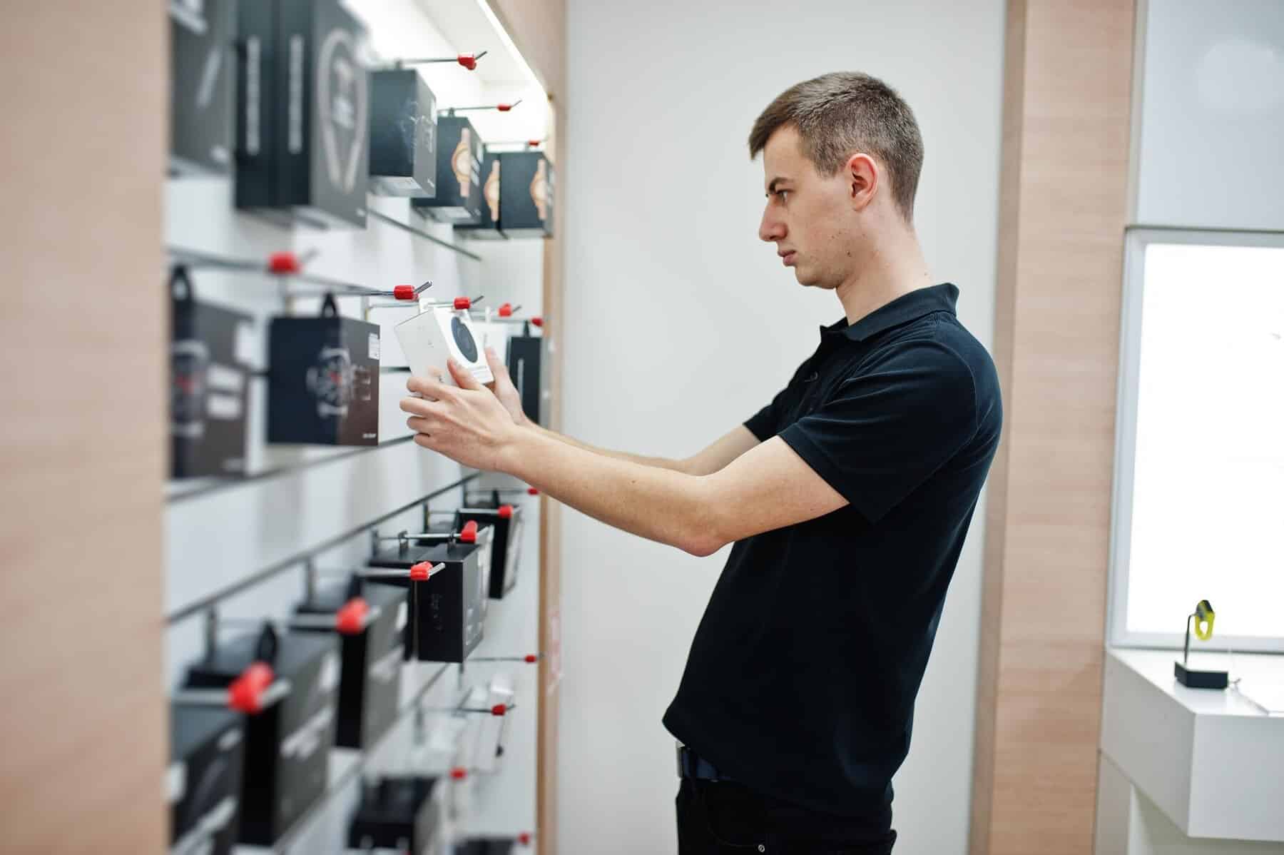 A person in a black polo shirt inspects a white electronic device while standing in a store showcasing various gadgets on shelves.