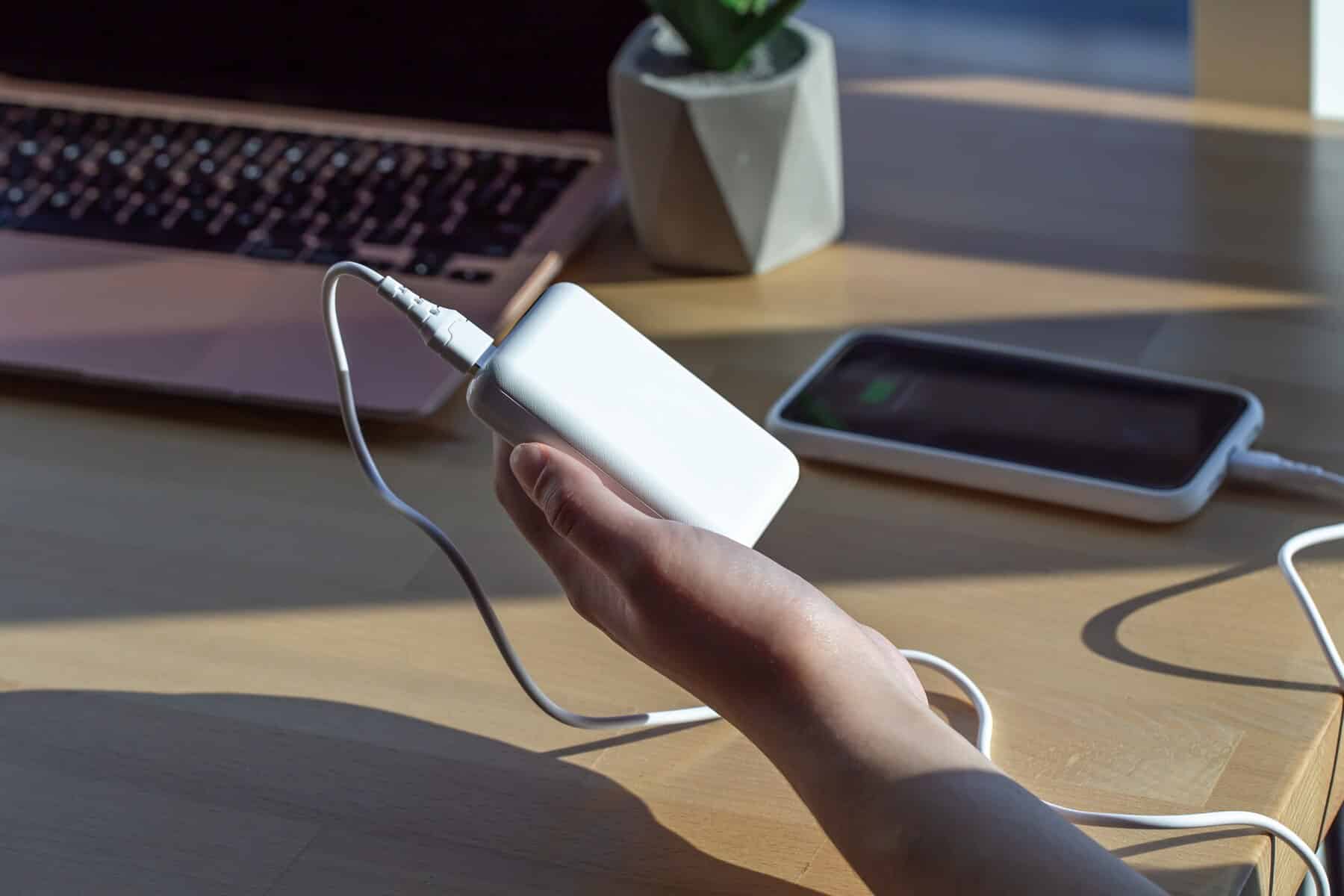 A person holding a portable battery pack connected to a charging cable, with a smartphone charging on a wooden desk beside a laptop and a small potted plant in a modern workspace.