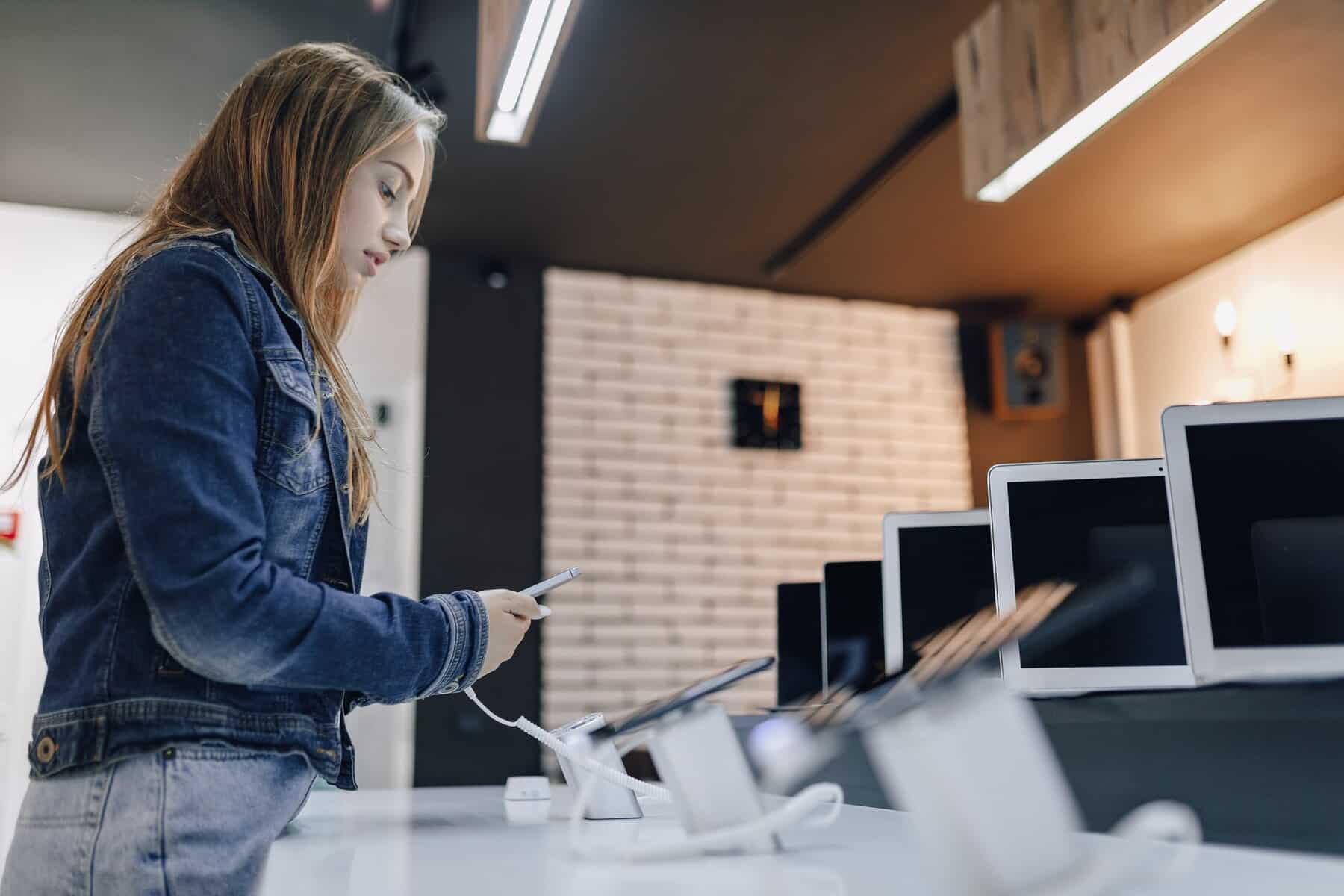 A young woman in a denim jacket examines a tablet at a tech store with several electronic devices displayed in front of her.