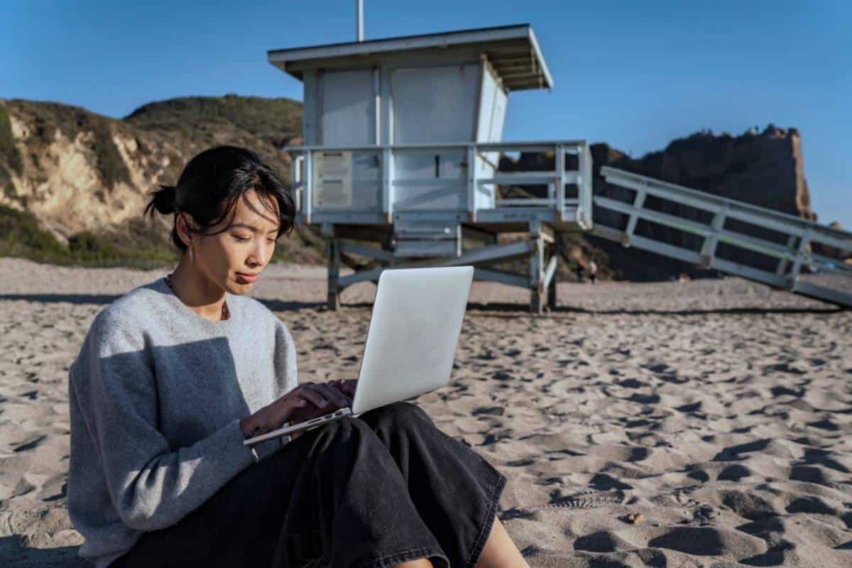 A person sits on the sandy beach, working on a laptop with a lifeguard