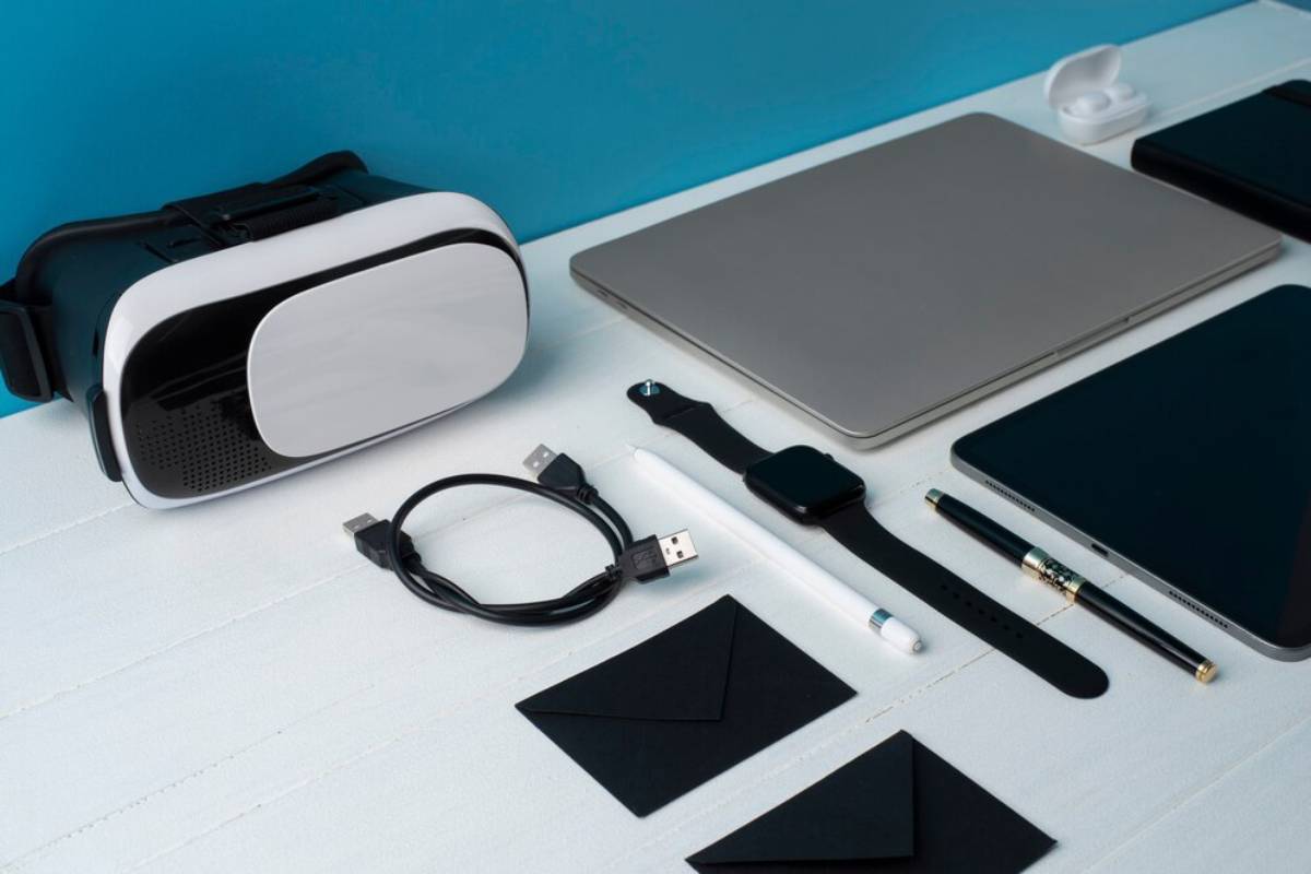 A VR headset, laptop, tablet, smartwatch, charging cable, stylus, black envelopes, and a pen arranged on a white surface.