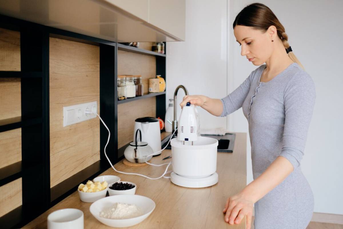 A person in a gray outfit uses a handheld mixer, surrounded by ingredients like flour, butter, and fruit on a sleek kitchen countertop.