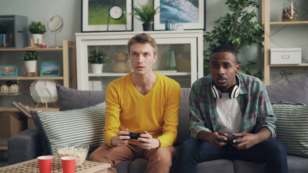 Two friends sit on a couch, focused on gaming with controllers in hand