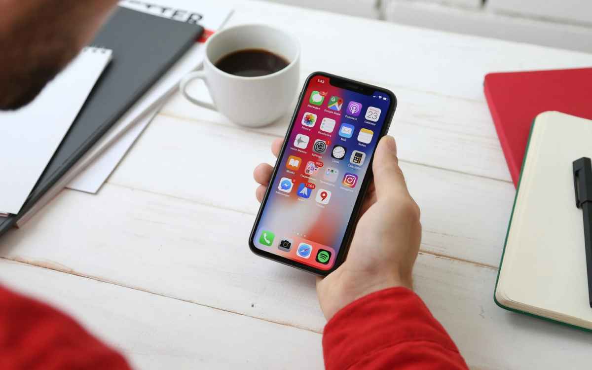 A person in a red shirt holds a smartphone displaying app icons, next to a cup of coffee and notebooks on a white desk.