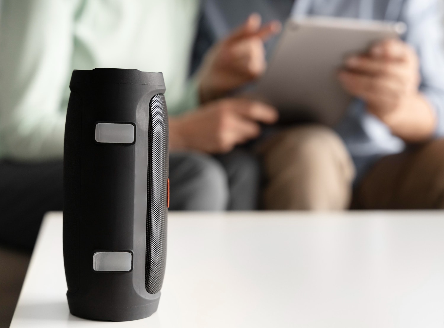 A portable black speaker sits on a white table, while two people in the background engaged in conversation, one holding a tablet.