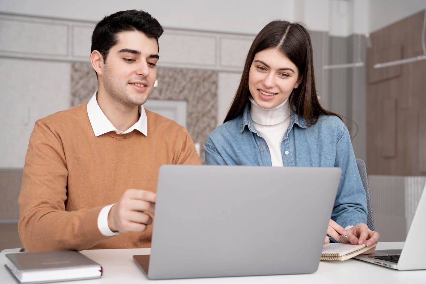 Two individuals collaborate at a desk, one gesturing towards a laptop while the other takes notes, set in a modern workspace.